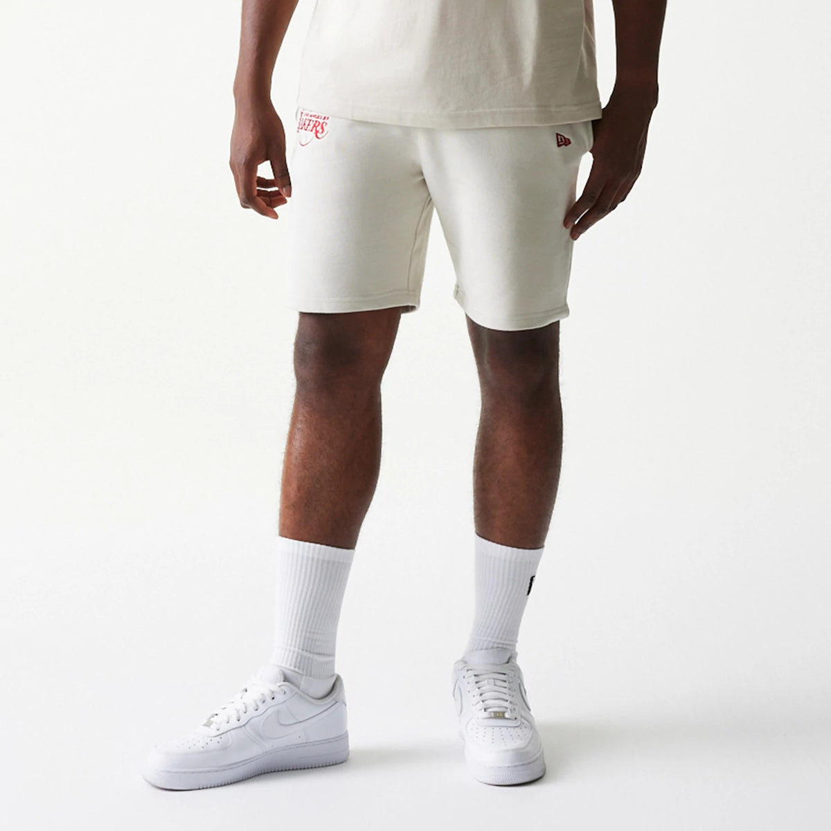 New Era LA Lakers NBA League Essential Cream Shorts, front view on-model showing the cream-coloured shorts with a red embroidered Lakers logo above the hem on the right leg and a small embroidered New Era flag logo on the left leg.