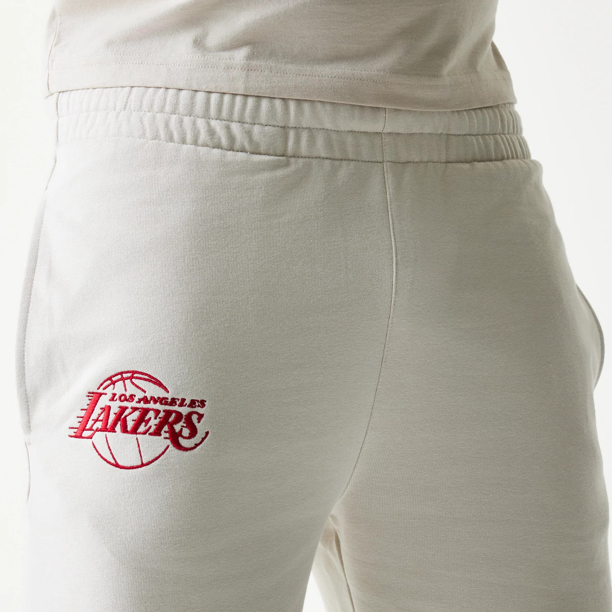 New Era LA Lakers NBA League Essential Cream Shorts, three-quarter view showing the elastic waistband and an embroidered red Lakers logo on the left thigh, worn with a matching cream top.