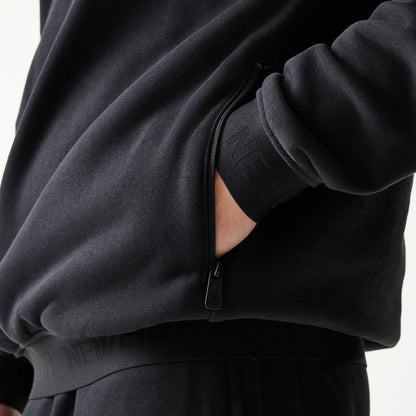New Era Tech Strip Black Oversized Pullover Hoodie, side view showing a hand in the front pocket with a zip closure, debossed New Era logo on the ribbed cuff, and another debossed logo on the hem.