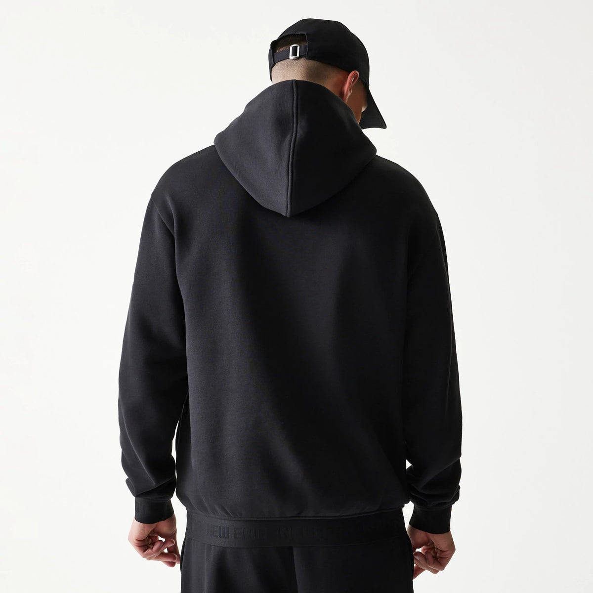 New Era Tech Strip Black Oversized Pullover Hoodie, back view on-model showing the plain black back with a large hood, New Era branding along the hem, styled with matching black trousers and a black cap worn by the model.