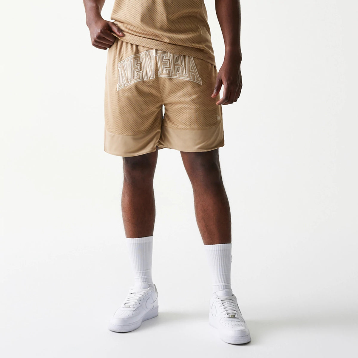 New Era Mesh Graphic Light Beige Oversized Shorts, front view showing large embroidered New Era lettering on the upper front, mesh texture throughout, and a small New Era logo on the lower left leg, styled with a matching top.