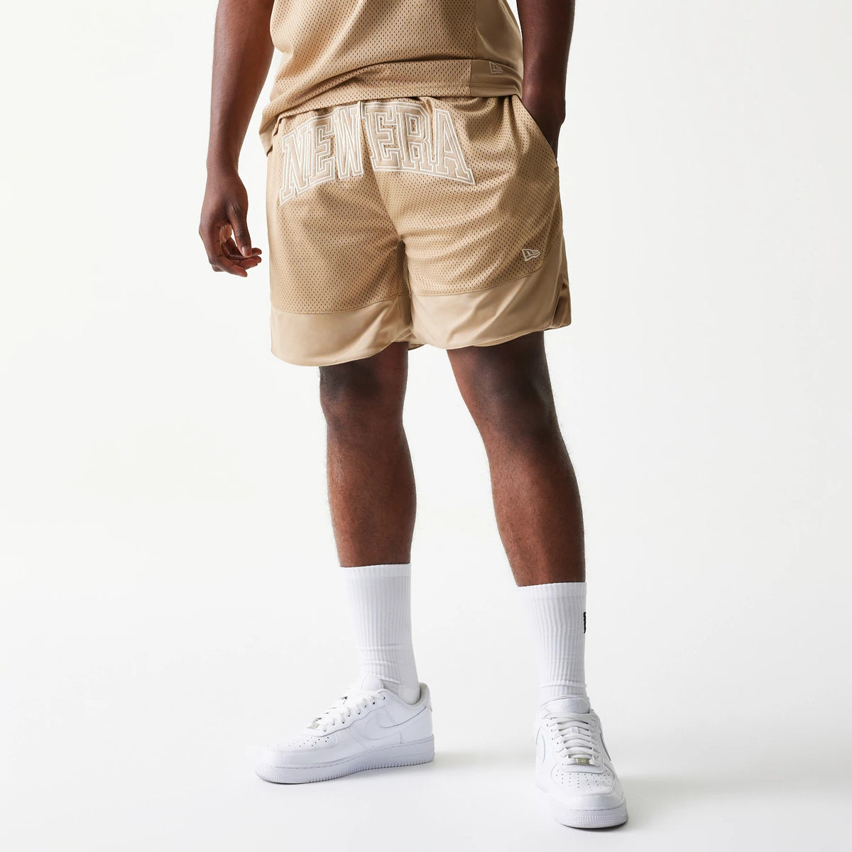 New Era Mesh Graphic Light Beige Oversized Shorts, back view on-model showing oversized white embroidered NEW ERA lettering across the back, the light beige mesh fabric, and the embroidered New Era logo near the hem.