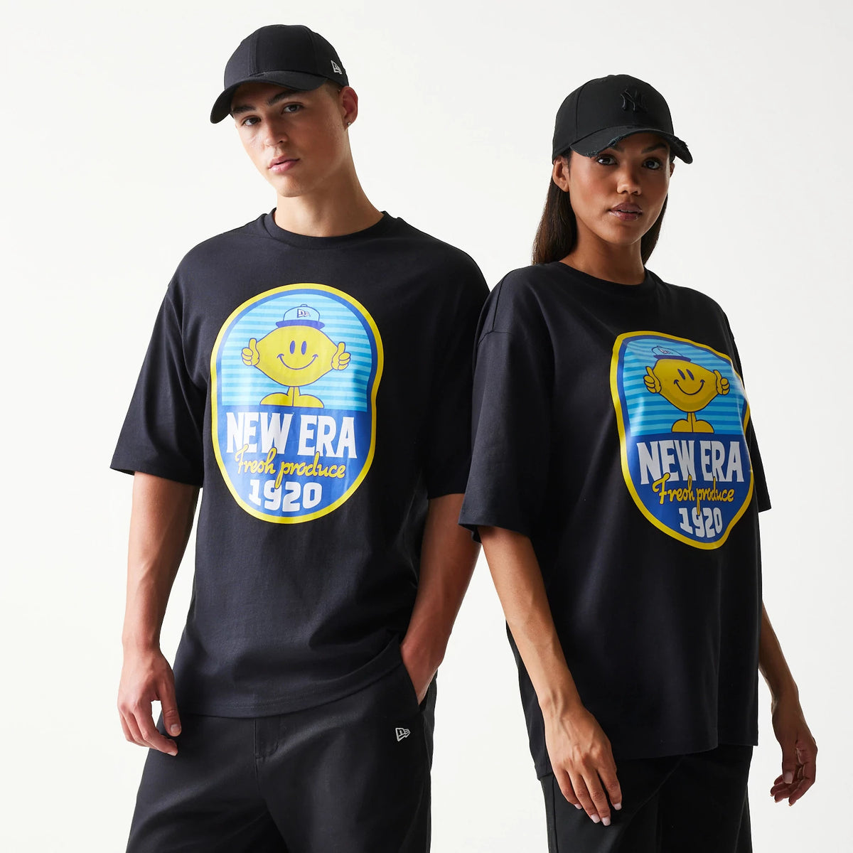 New Era Lemon Fruit Graphic Black Oversized T-Shirt, front view on models styled with black caps, showing a large graphic of a smiling lemon character with a blue cap and text reading New Era fresh produce 1920 on the chest.