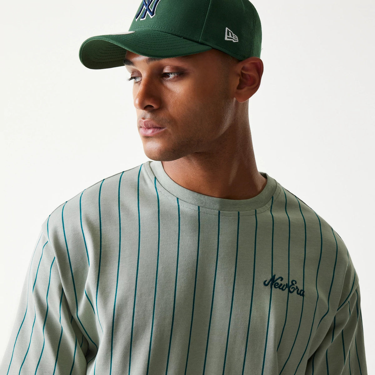 New Era Pinstripe Pastel Green Oversized T-Shirt, on-model front view showing vertical dark green pinstripes on the pastel green fabric and an embroidered New Era logo on the left chest, styled with a matching green cap.