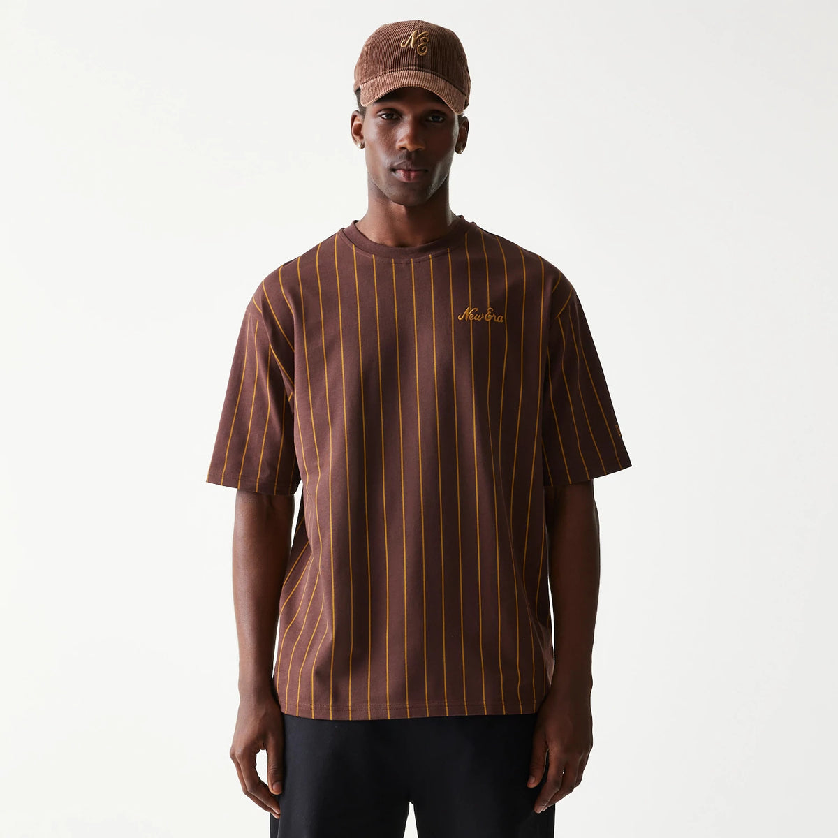 New Era Pinstripe Brown Oversized T-Shirt, front view on-model showing the brown tee with vertical yellow pinstripes, an embroidered New Era logo on the chest, styled with a matching brown cap featuring an embroidered logo.