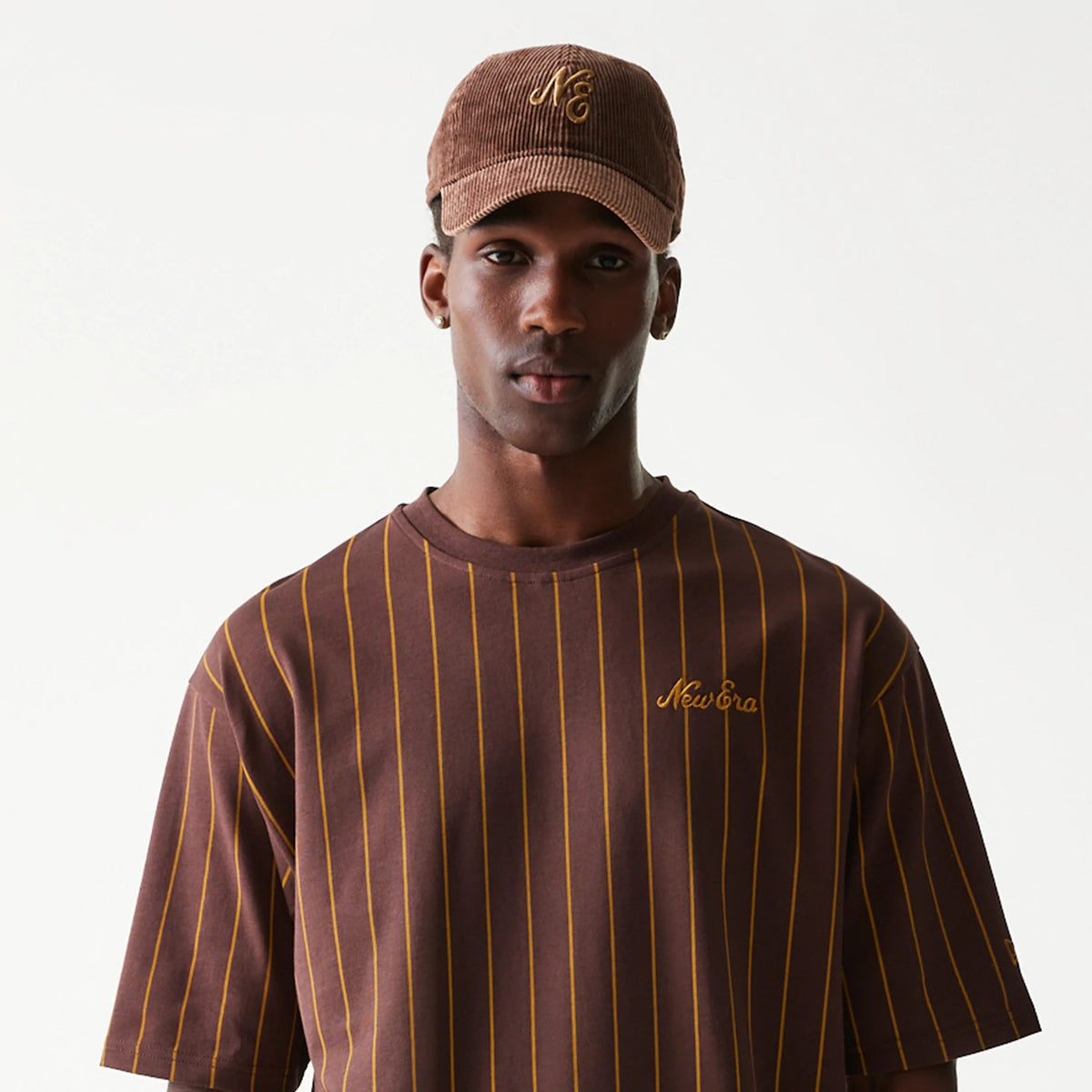 New Era Pinstripe Brown Oversized T-Shirt, front view on a model styled with a matching cap, featuring thin yellow pinstripes across the brown fabric and an embroidered New Era logo on the left chest.