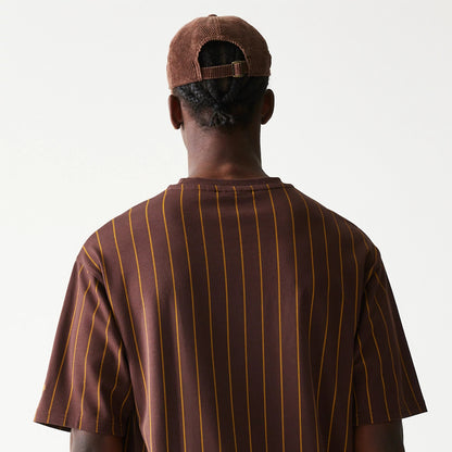 New Era Pinstripe Brown Oversized T-Shirt, back view showing vertical mustard yellow pinstripes running down the dark brown fabric, worn by a model with a brown cap, large loose sleeves and a relaxed fit.