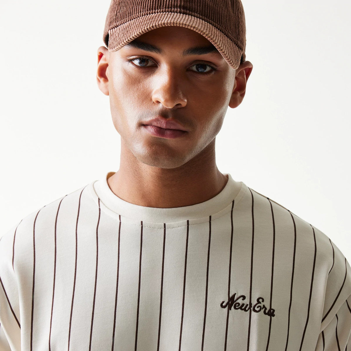 New Era Pinstripe Light Beige Oversized T-Shirt, front view showcasing thin vertical pinstripes and an embroidered New Era logo on the left chest, worn under a brown cap.