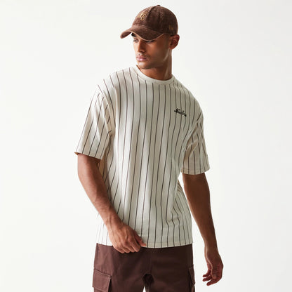 New Era Pinstripe Light Beige Oversized T-Shirt, front view on a model wearing a brown cap, featuring vertical thin black stripes and an embroidered New Era logo on the left chest.