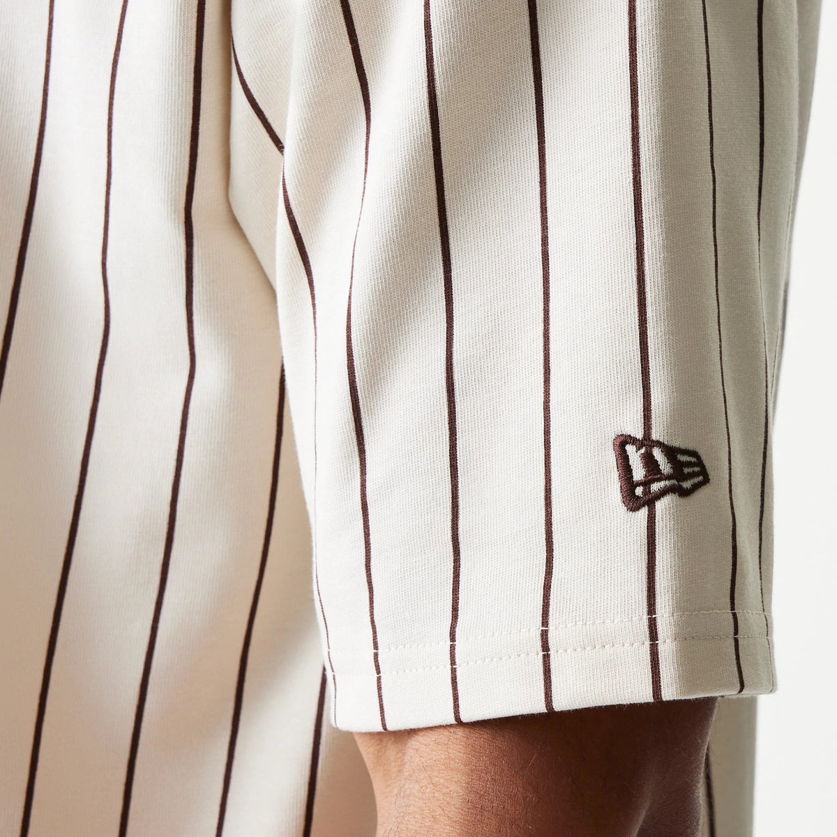New Era Pinstripe Light Beige Oversized T-Shirt, three-quarter view showing a close-up of dark pinstripes running vertically down the light beige fabric, with a dark brown embroidered New Era logo on the sleeve cuff.