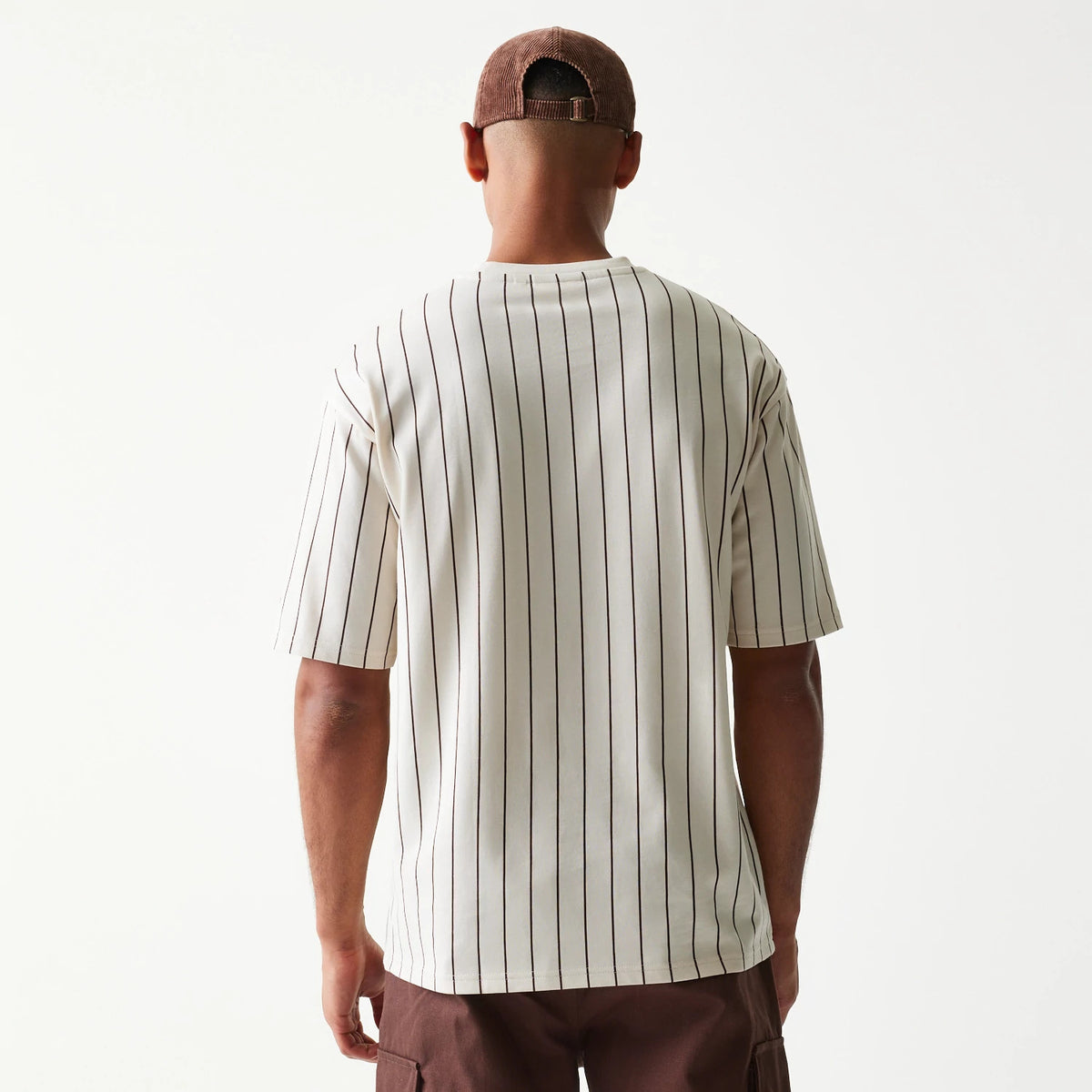 New Era Pinstripe Light Beige Oversized T-Shirt, back view showing vertical pinstripes running uniformly down the light beige fabric, worn by a model with a matching cap, highlighting the relaxed, oversized fit and short sleeves.