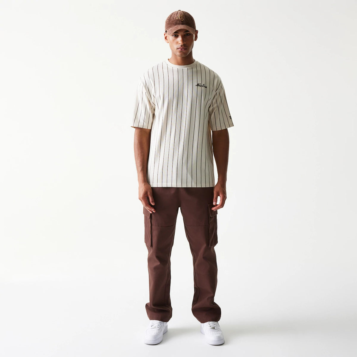 New Era Pinstripe Light Beige Oversized T-Shirt, front view on a model paired with brown trousers and a cap, featuring vertical black pinstripes and an embroidered New Era logo on the chest.