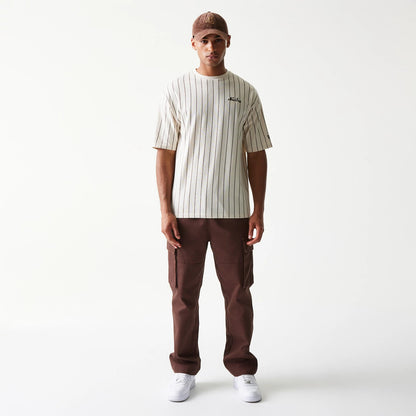 New Era Pinstripe Light Beige Oversized T-Shirt, front view on a model paired with brown trousers and a cap, featuring vertical black pinstripes and an embroidered New Era logo on the chest.