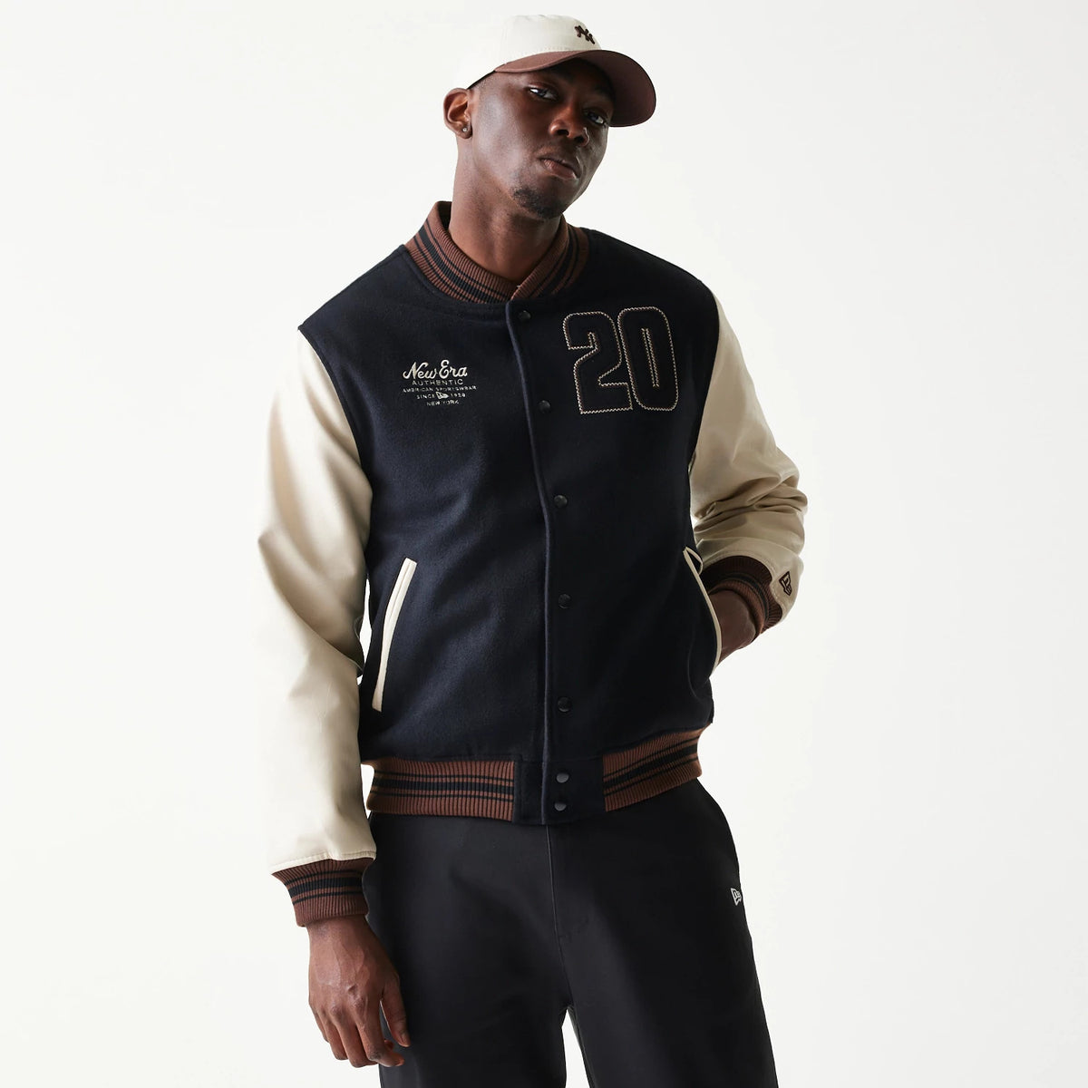 New Era Varsity Black Jacket, front view on-model showing a black body with cream sleeves, contrasting ribbed cuffs, and waistband, embroidered New Era logo on the chest, number 20 applique on the right side, paired with a cream cap.