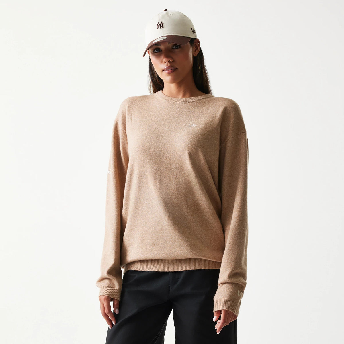 New Era Essential Flag Light Beige Knitted Sweater, front view on a model with a New York Yankees cap, showing the small white embroidered New Era logo on the left chest and right sleeve cuff.
