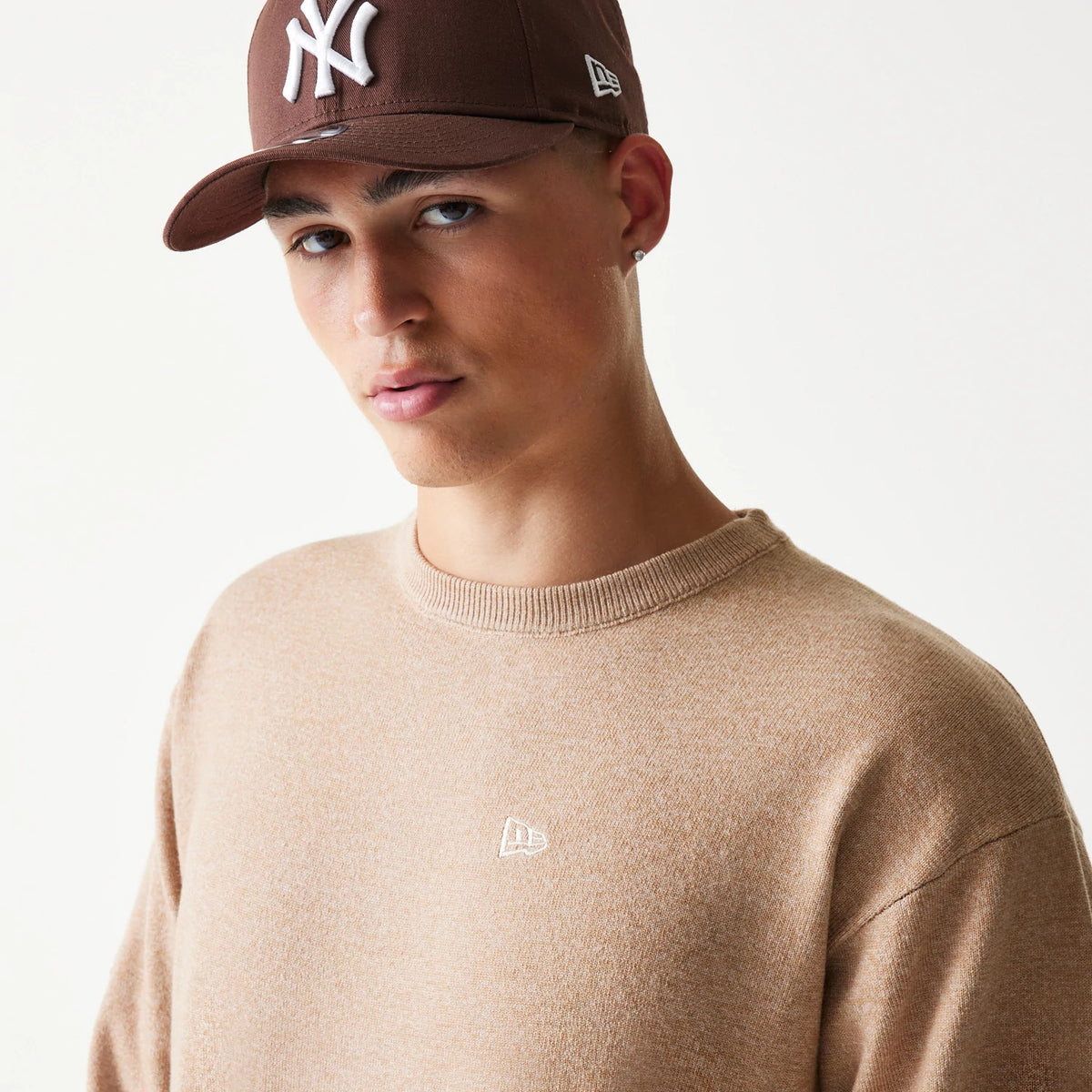 New Era Essential Flag Light Beige Knitted Sweater, on-model view featuring a brown New York Yankees 59FIFTY cap, showing the embroidered New Era flag logo on the chest and a ribbed crew neckline.