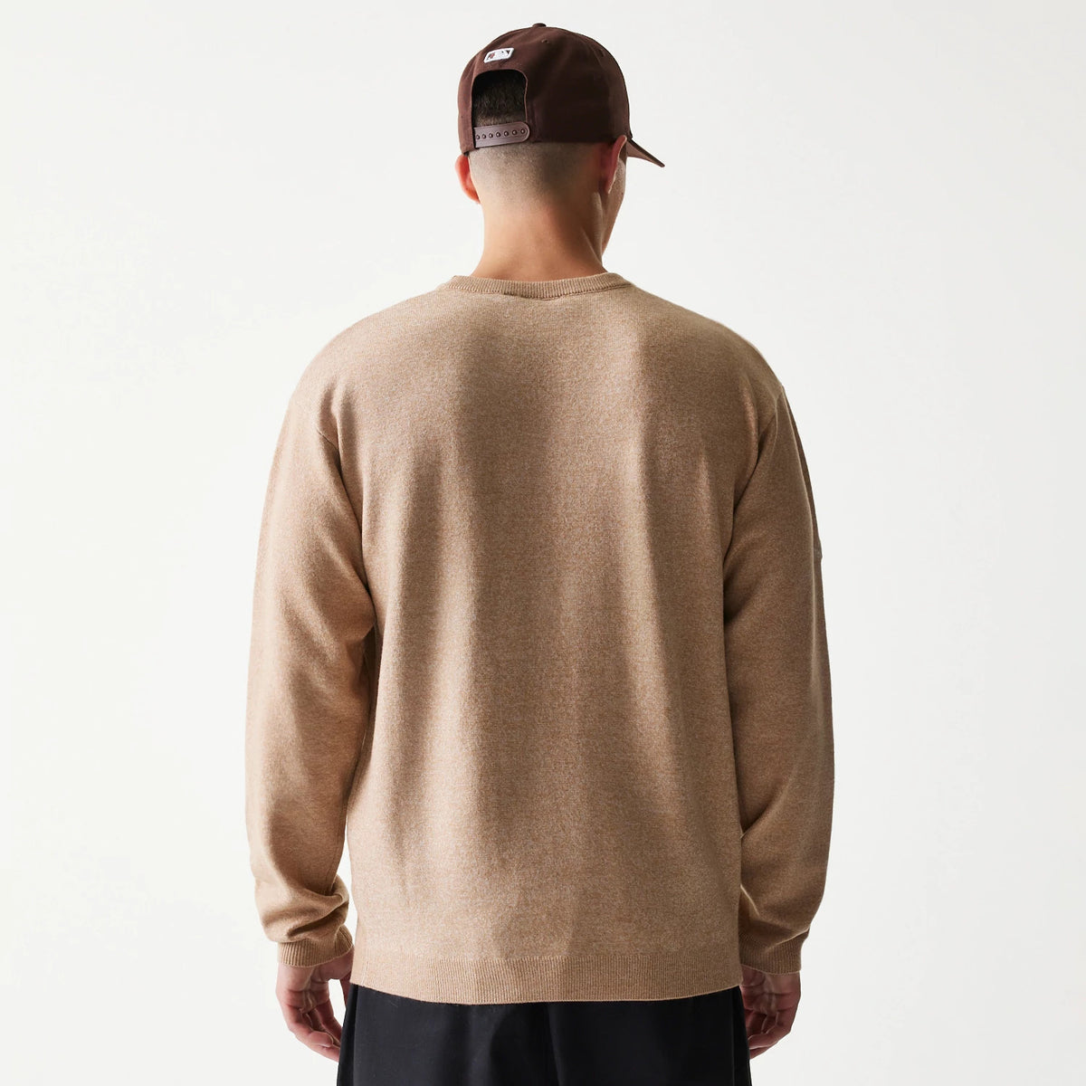 New Era Essential Flag Light Beige Knitted Sweater, back view on a model wearing a dark cap, showing the light beige texture of the sweater with ribbed cuffs and hem, offering a relaxed fit over dark trousers.