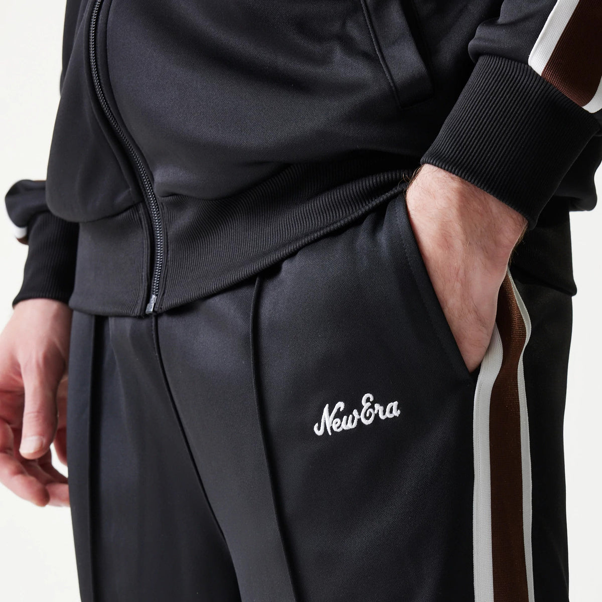 New Era Script Black Straight Leg Joggers, close-up view showing white New Era script logo embroidered on the right thigh, with brown and white side stripes and a hand in the pocket.