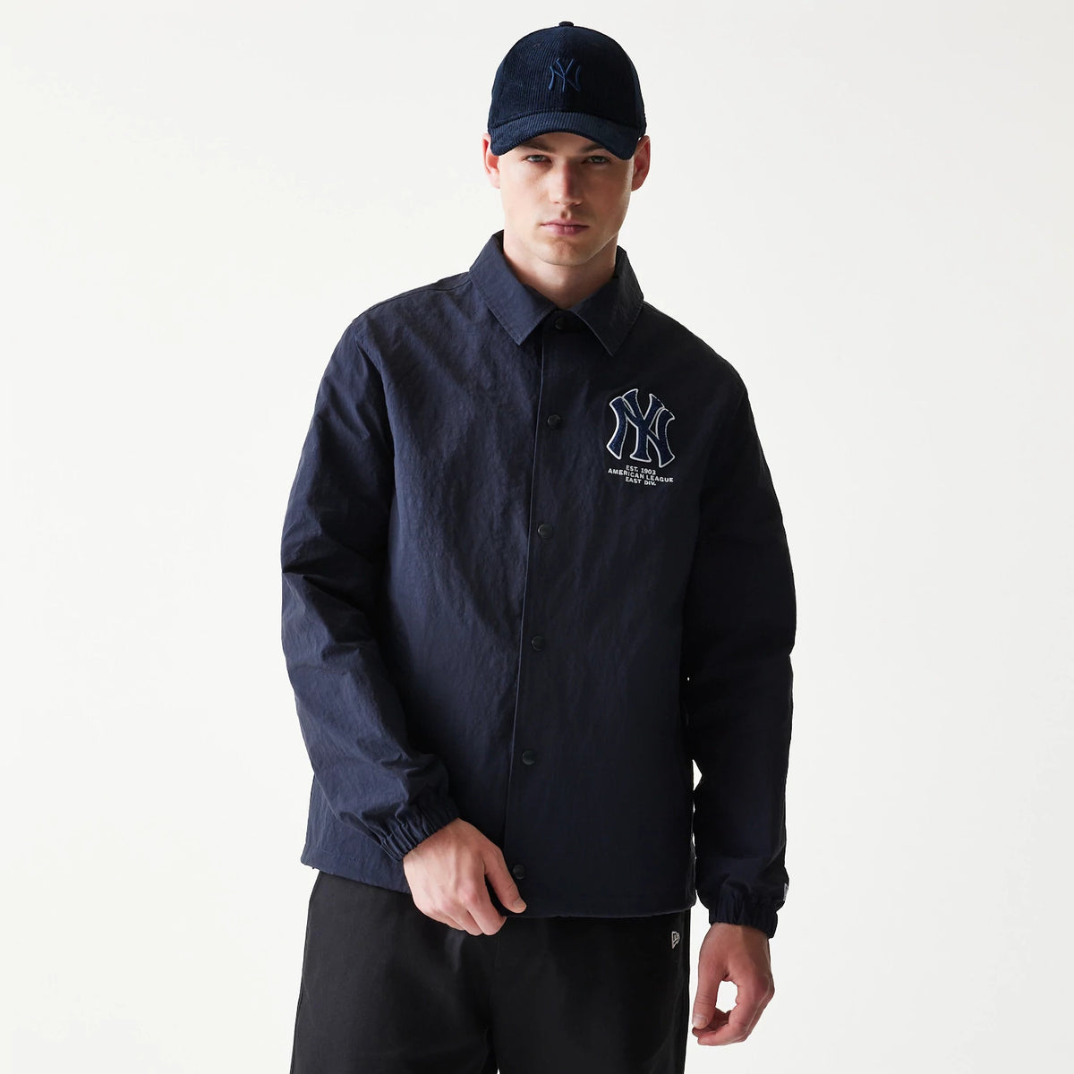 New Era New York Yankees MLB Coaches Navy Jacket, front view on model showing the embroidered Yankees logo on the left chest, a button-up front with a pointed collar, and elasticated cuffs, paired with a matching navy cap featuring a raised logo.