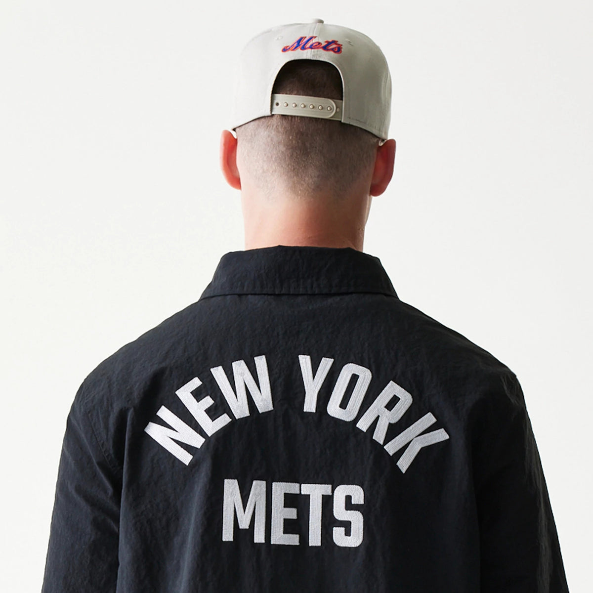 New Era New York Mets MLB Coaches Black Jacket, back view on a model showing white New York Mets lettering across the upper back, styled with a grey cap featuring a Mets logo embroidered above the adjustable strap.