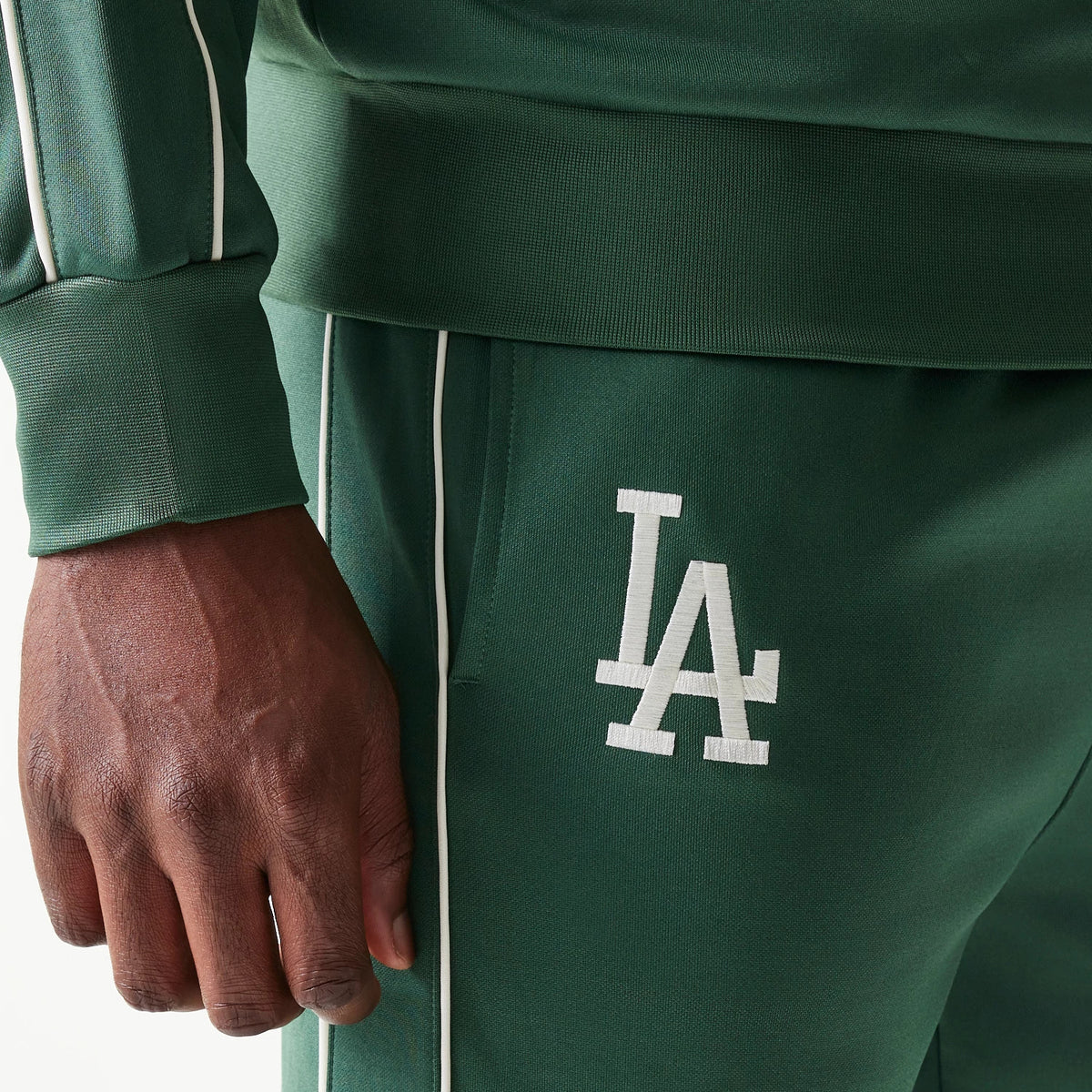 New Era LA Dodgers MLB Piping Dark Green Straight Leg Joggers, close-up showing the white embroidered LA logo on the thigh, with thin white pipe running down the side seams and an elasticated waistband.