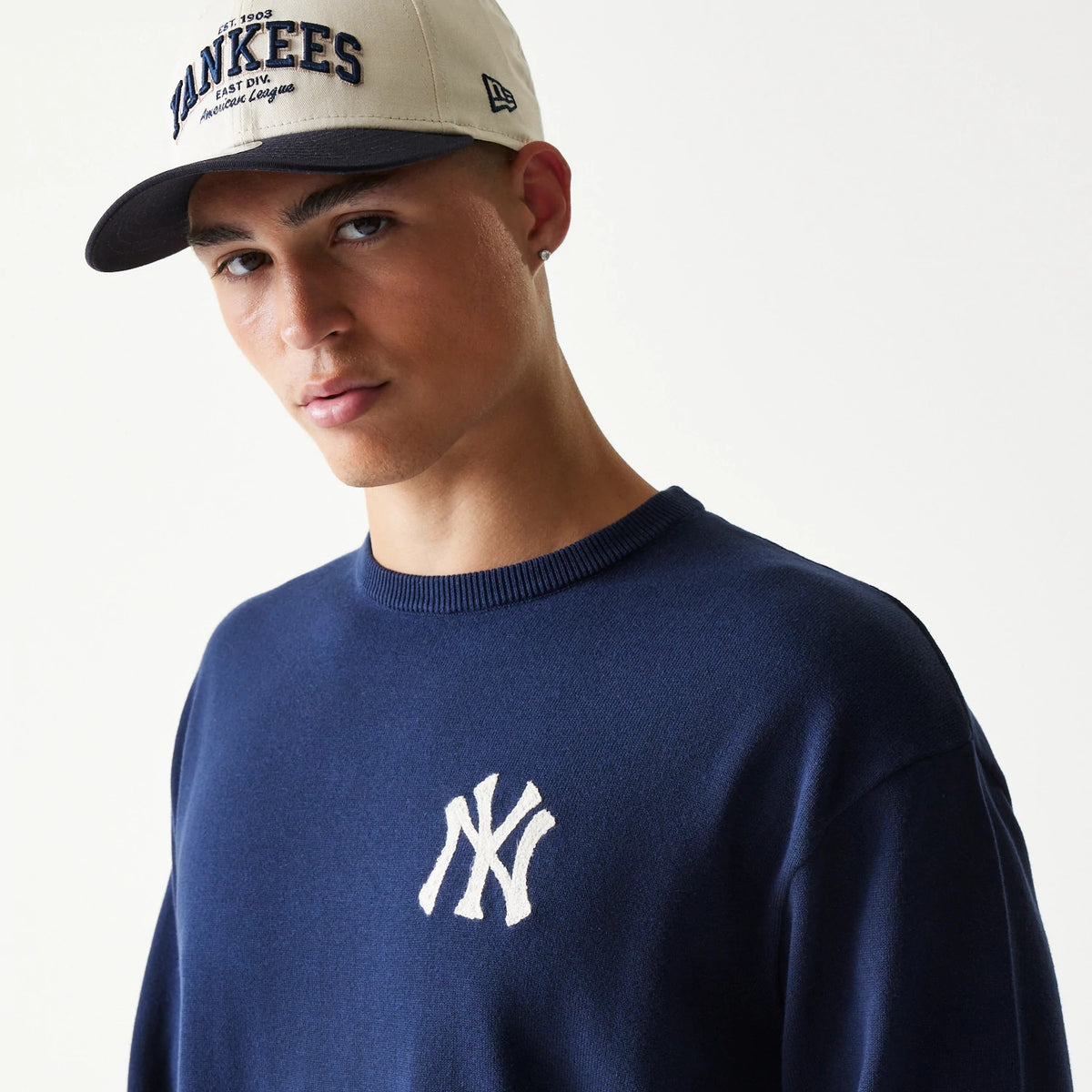 New Era New York Yankees MLB Essential Navy Knitted Sweater, on-model view showing the white embroidered Yankees logo on the chest, worn by a model also wearing a beige Yankees cap with navy visor and New Era flag logo on the side.