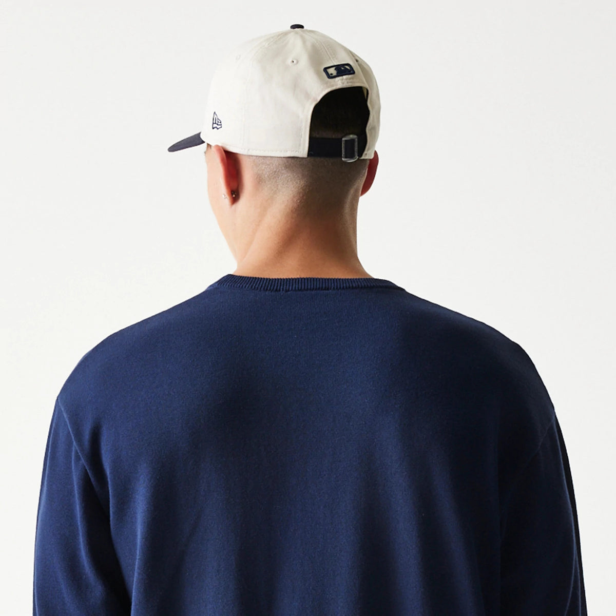 New Era New York Yankees MLB Essential Navy Knitted Sweater, rear view on a model also wearing a light-coloured adjustable cap, showing the plain back of the sweater with a ribbed neckline and the MLB logo on the cap's back strap.