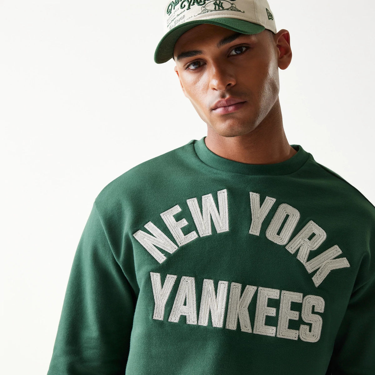 New Era New York Yankees MLB Graphic Dark Green Oversized Crew Neck Sweater, front view on a model with white and grey textured lettering displaying the team's name across the chest, styled with a light cap.