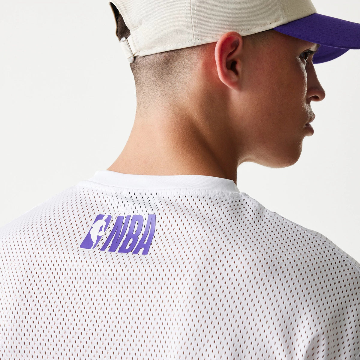 New Era LA Lakers NBA Graphic Mesh White T-Shirt, back view on-model showing the white mesh fabric with purple NBA lettering just below the neckline, paired with a beige and purple cap worn by the model.