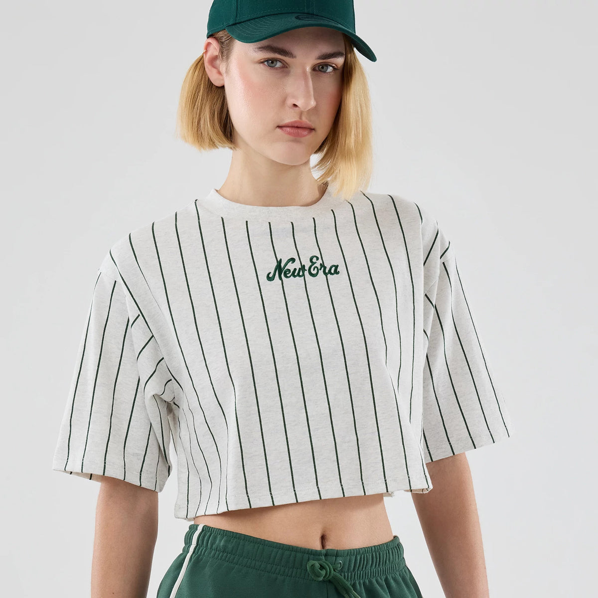 Womens New Era Pinstripe Pastel Grey Cropped T-shirt, front view on-model showing vertical dark green pinstripes on a grey background with an embroidered New Era logo at the centre of the chest, paired with matching green sports cap and trousers.