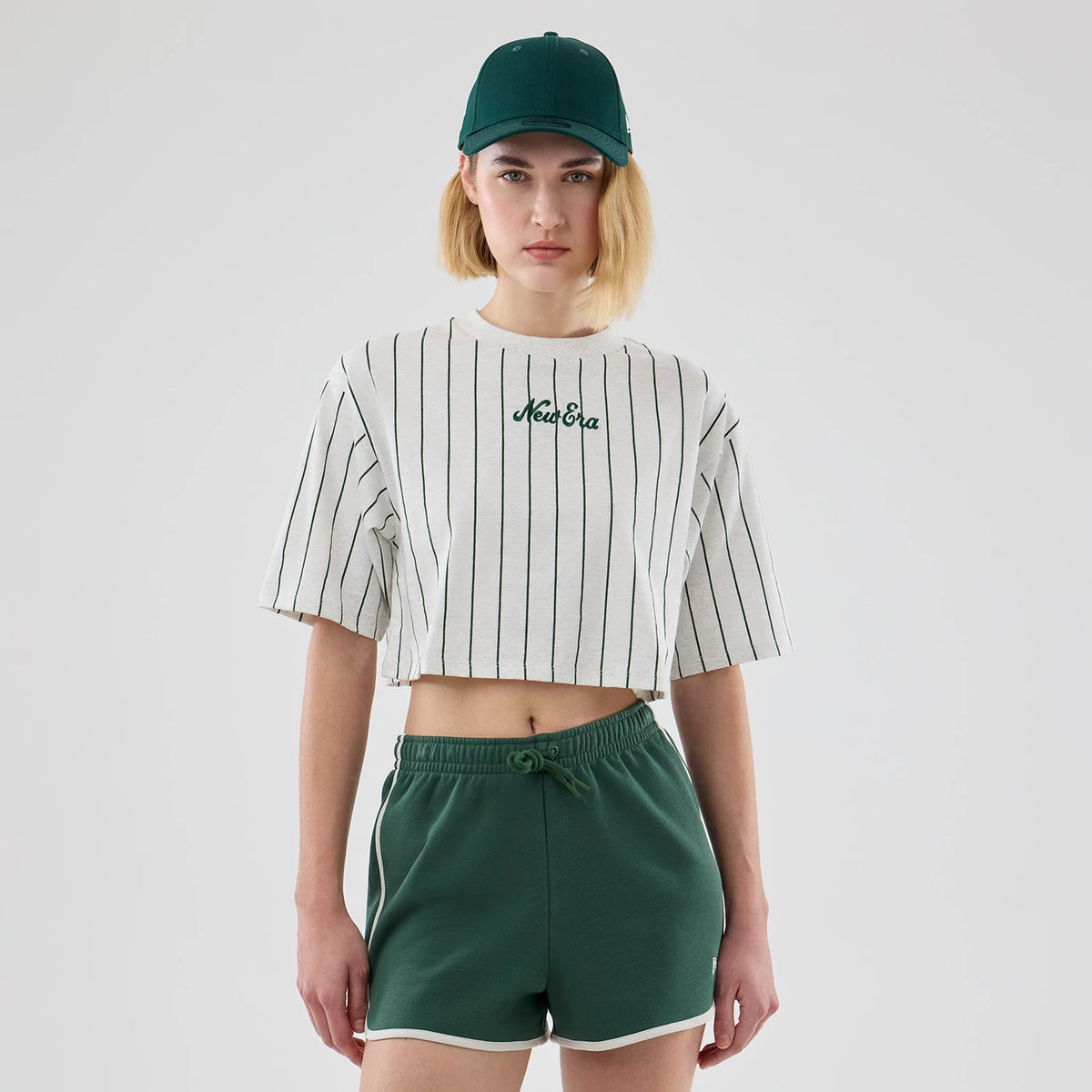 Womens New Era Pinstripe Pastel Grey Cropped T-shirt, front view on a model styled with green shorts and a dark green cap, featuring thin black pinstripes and an embroidered New Era logo centred on the chest.