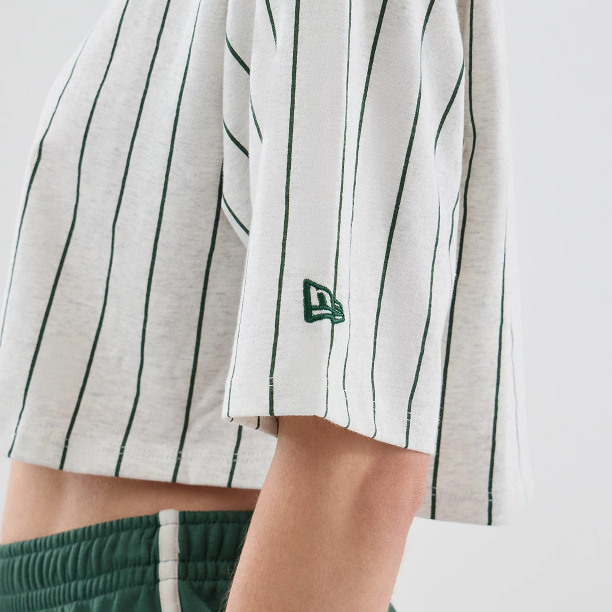 Womens New Era Pinstripe Pastel Grey Cropped T-shirt, side view showing vertical dark green pinstripes along the light grey fabric and an embroidered green New Era logo on the lower sleeve, styled with dark green shorts.