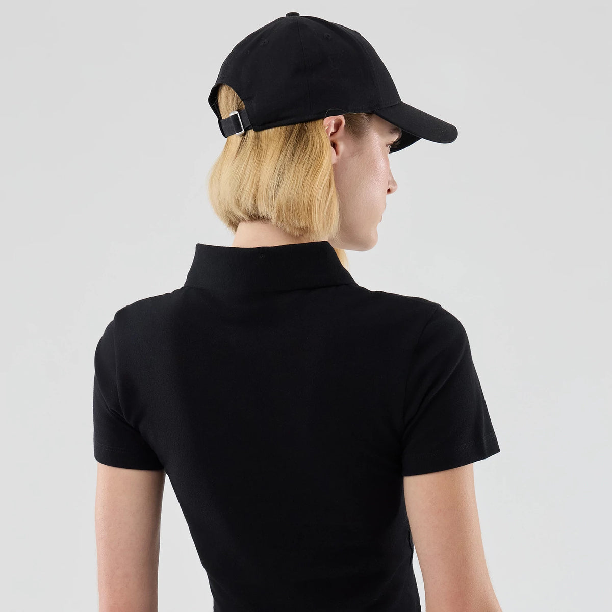 New Era New York Yankees Womens MLB Black Cropped Slim Polo T-shirt, back three-quarter view on-model showing the black fabric with a folded collar, short sleeves, and styled with a matching black cap featuring a buckle at the back.