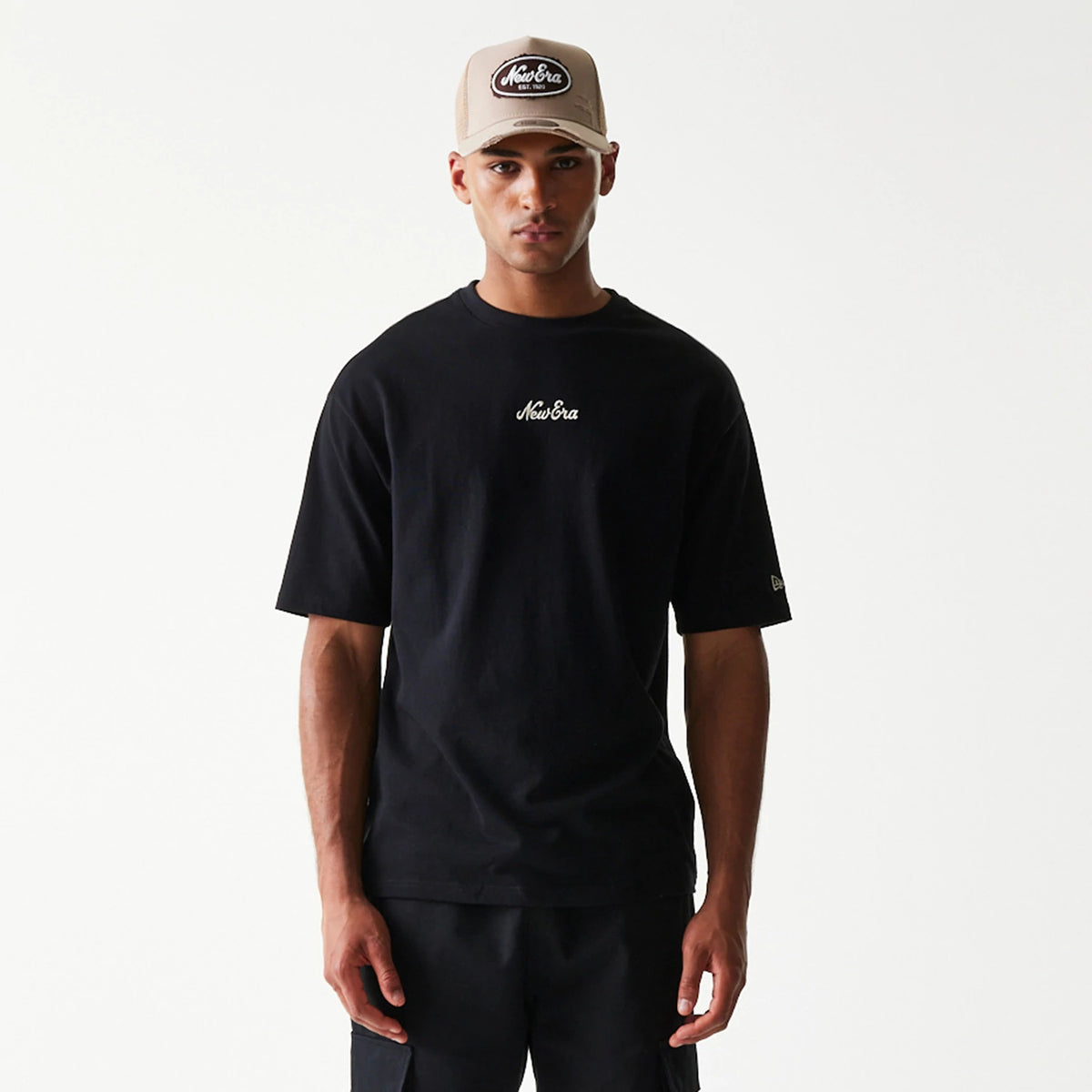 New Era Heritage Script Black T-Shirt, front view on-model showing the embroidered New Era script logo in white on the chest and a small embroidered New Era logo on the left sleeve, styled with a beige New Era cap.