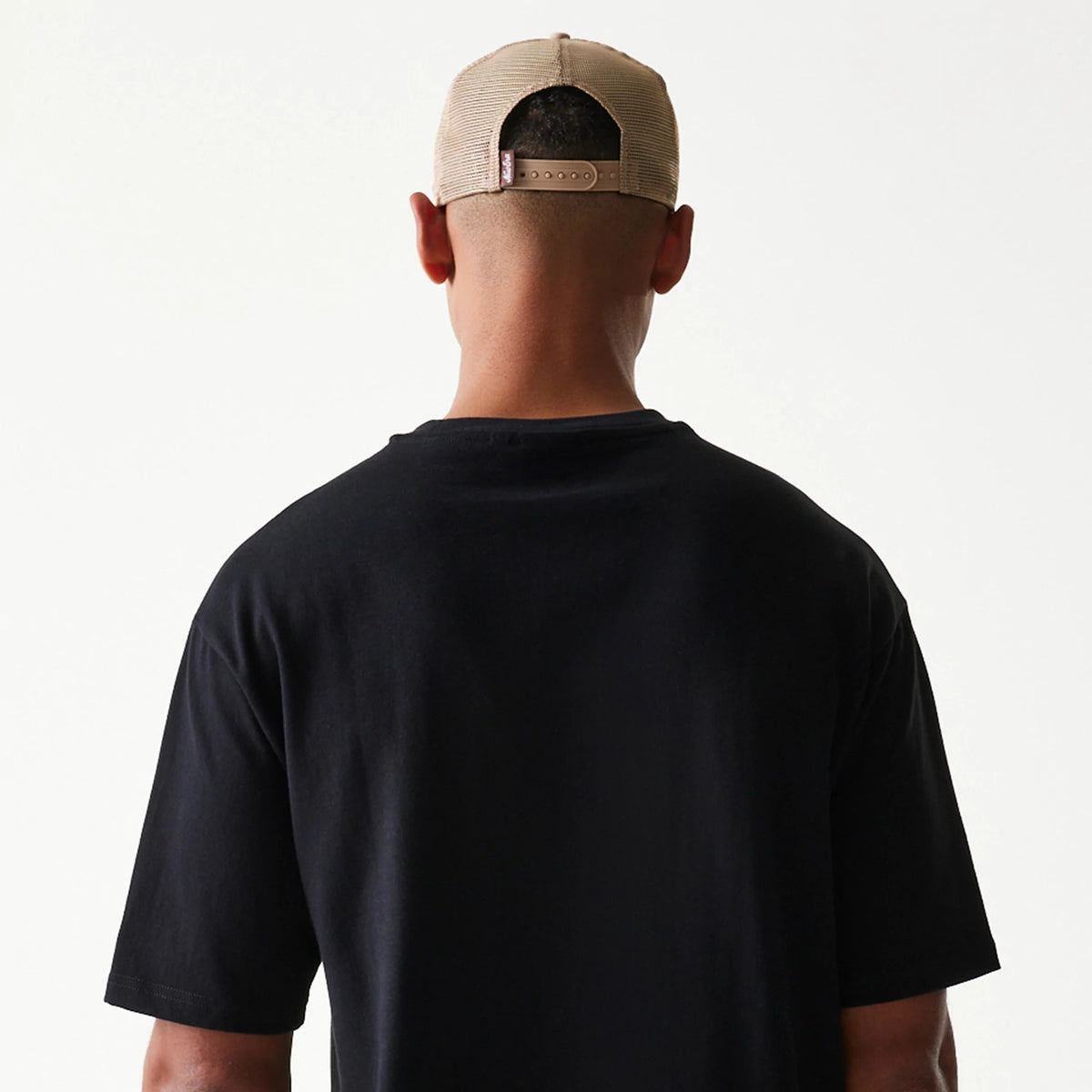 New Era Heritage Script Black T-Shirt, back view on a model with a beige cap, showing the plain black fabric of the T-shirt with short sleeves and a relaxed fit extending over the shoulders.