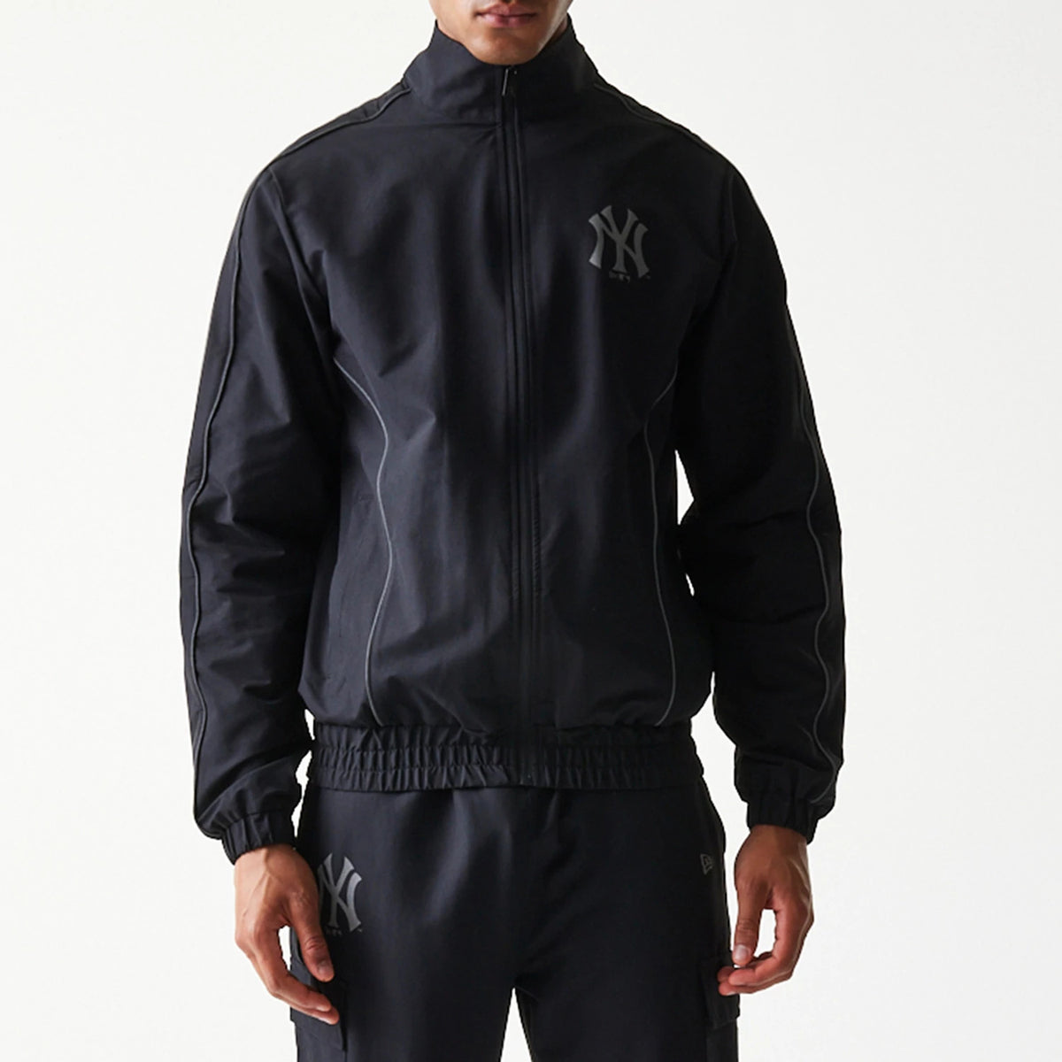 New Era New York Yankees MLB Black Track Jacket, front view on-model showing the full zip closure, raised NY logo on the left chest, high collar and elasticated cuffs, styled with matching black trousers featuring a similar NY logo.