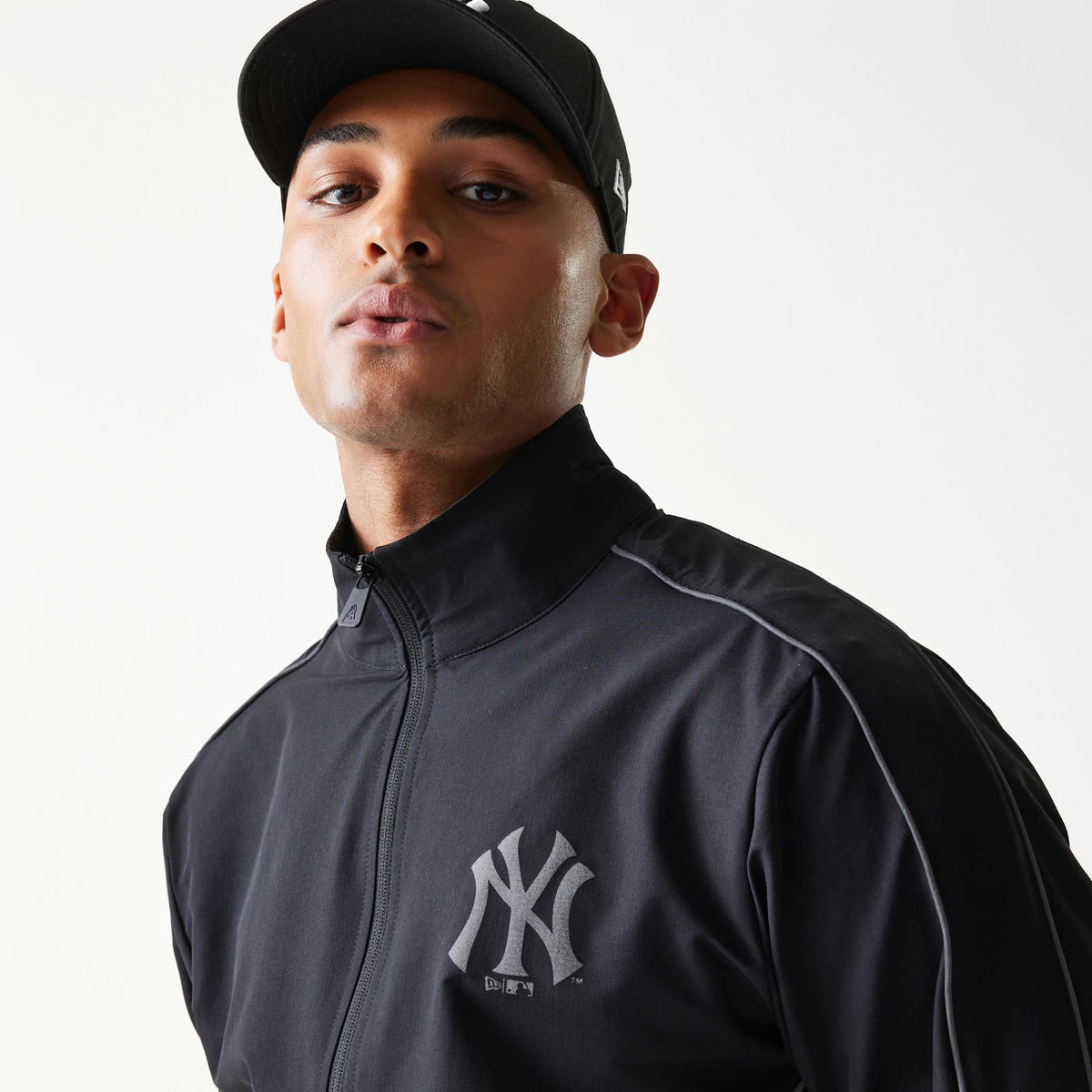 New Era New York Yankees MLB Black Track Jacket, front view on a model highlighting the raised New York Yankees logo on the left chest and a zip closure, paired with a matching black cap featuring an embroidered logo.