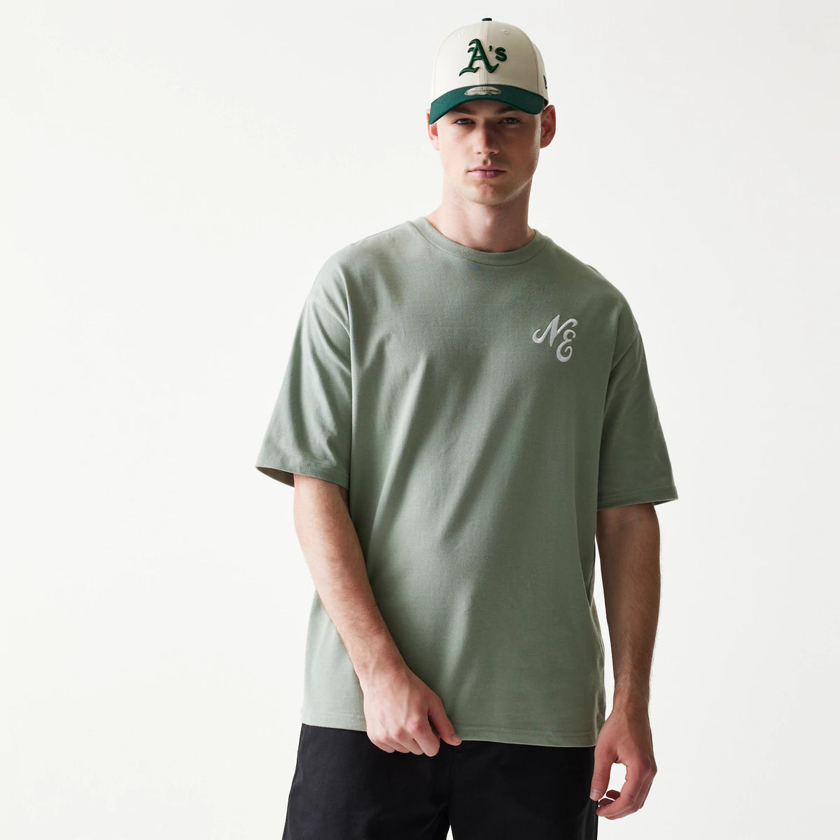 New Era Floral Graphic Pastel Green Oversized T-Shirt, front view on-model displaying a minimalist design with a white embroidered NE logo on the upper left chest, paired with black trousers and a green and white cap.