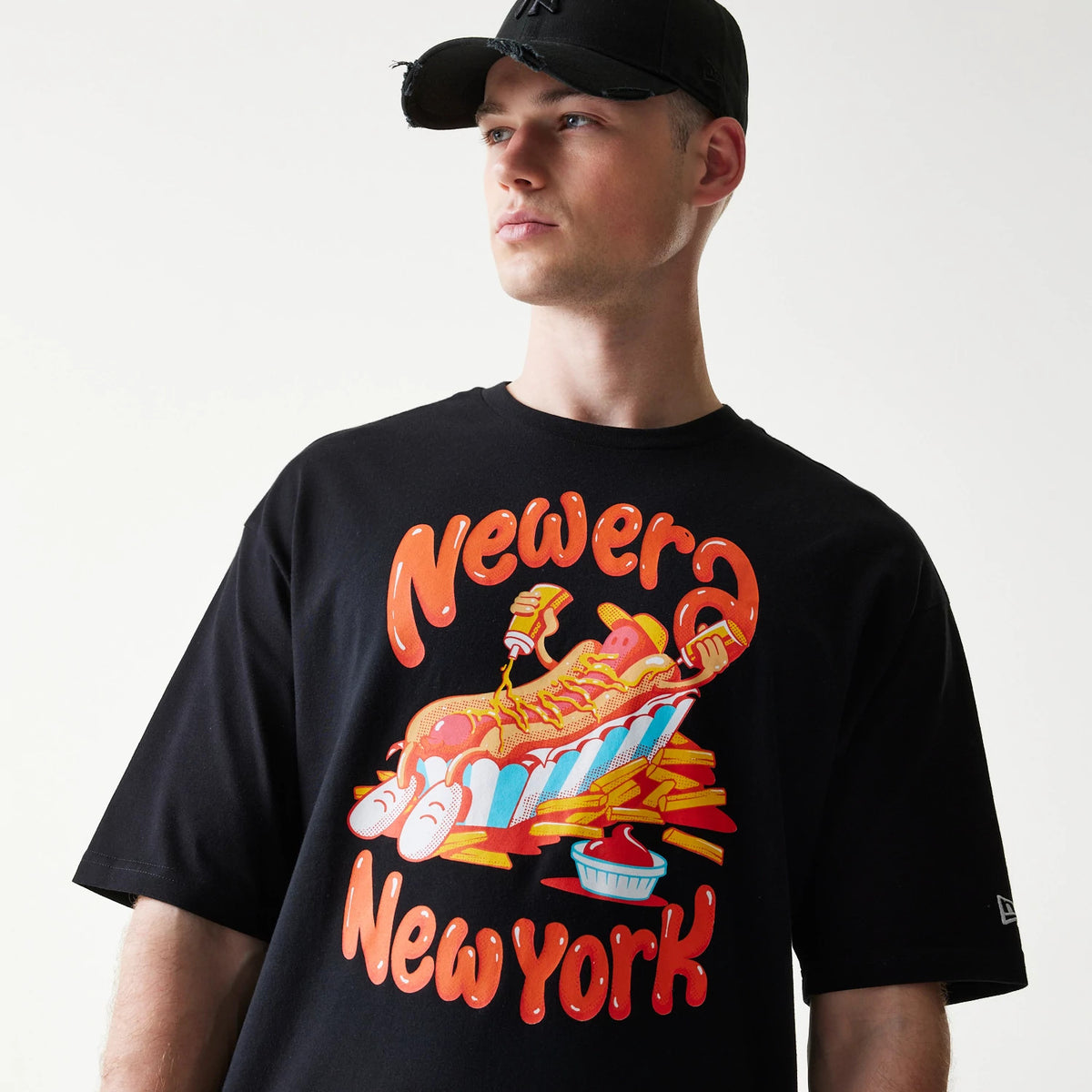 New Era Hotdog Food Graphic Black Oversized T-Shirt, front view on-model showing a hotdog graphic with a playful cartoon figure and 'New Era New York' lettering on the black fabric, with a small embroidered New Era logo on the sleeve.