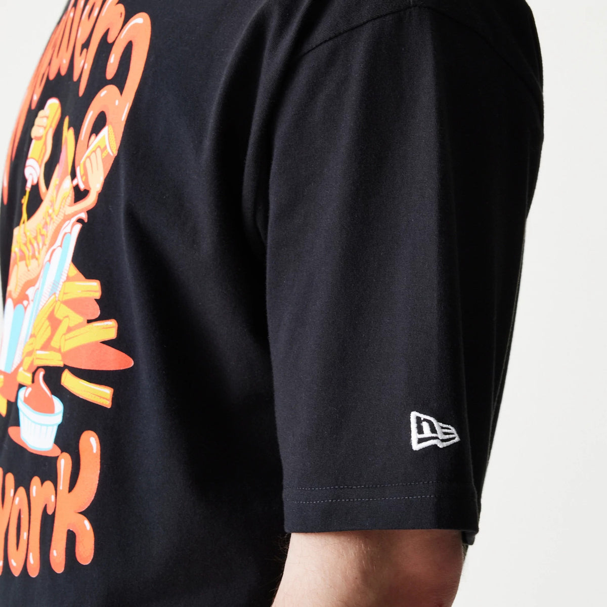 New Era Hotdog Food Graphic Black Oversized T-Shirt, side view showing a hotdog-themed graphic in orange and blue on the black fabric, and a white embroidered New Era logo on the sleeve near the cuff.