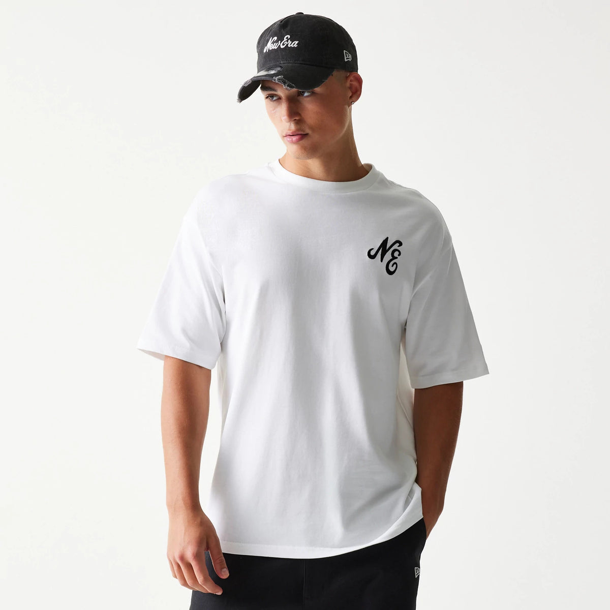 New Era Floral Graphic White Oversized T-Shirt, front view on a model showing the large white tee with a small black embroidered NE logo on the chest, styled casually with a matching black New Era cap.