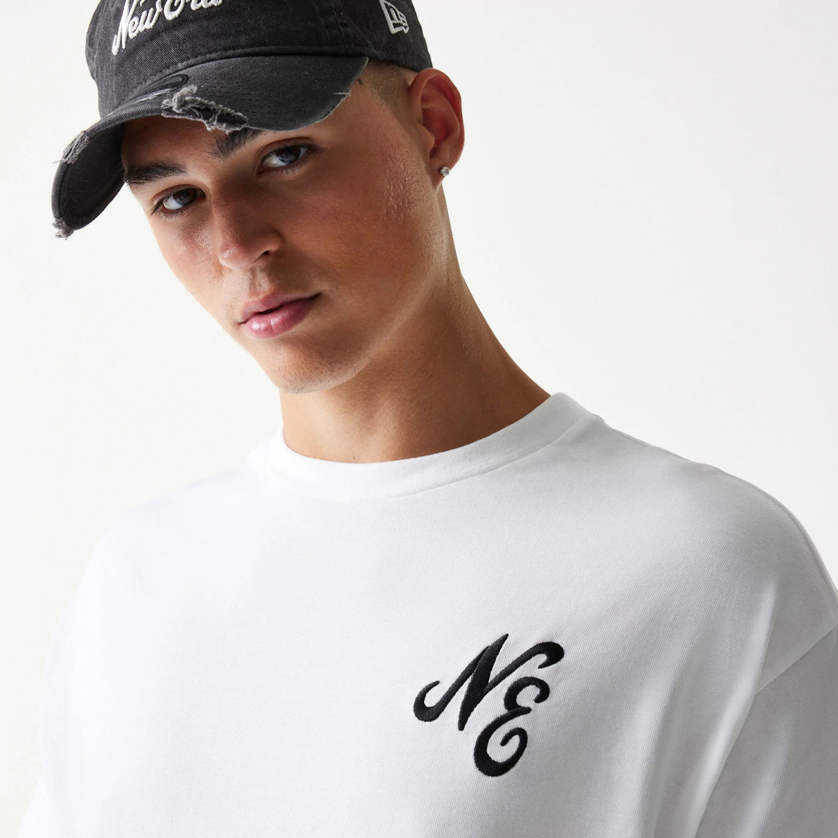 New Era Floral Graphic White Oversized T-Shirt, front view on-model showing the black embroidered NE logo on the chest, styled with a black distressed cap featuring a white embroidered New Era script and logo on the side.