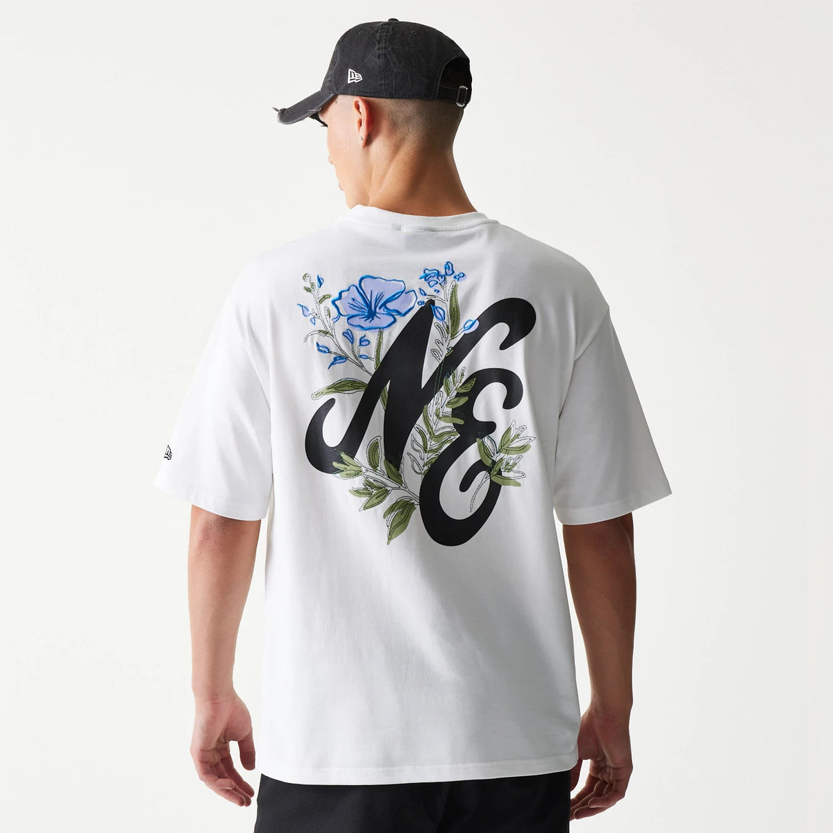 New Era Floral Graphic White Oversized T-Shirt, back view on a model with a black cap, featuring a large black NE graphic with blue flowers and green leaves printed across the upper back.