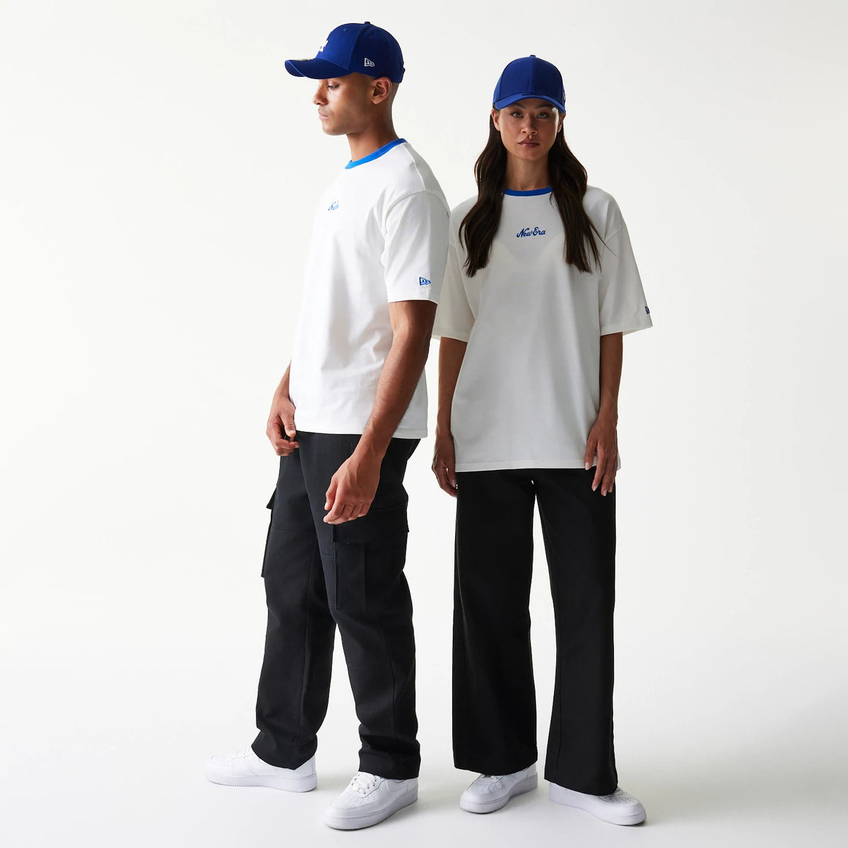 New Era Script White Oversized T-Shirt, front view on two models paired with blue caps, featuring the embroidered New Era script logo centred on the chest and a contrasting blue neckline accentuating the white fabric.