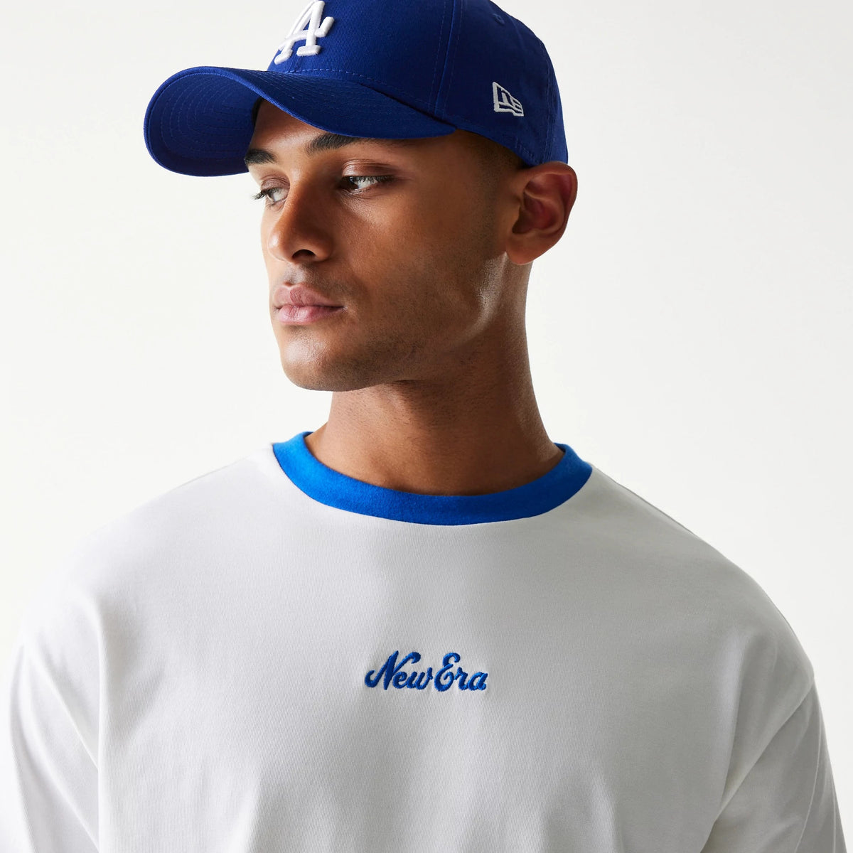New Era Script White Oversized T-Shirt, front view on-model showing a blue embroidered New Era logo on the chest and a blue neckline, worn with a blue cap featuring a white embroidered letter logo.