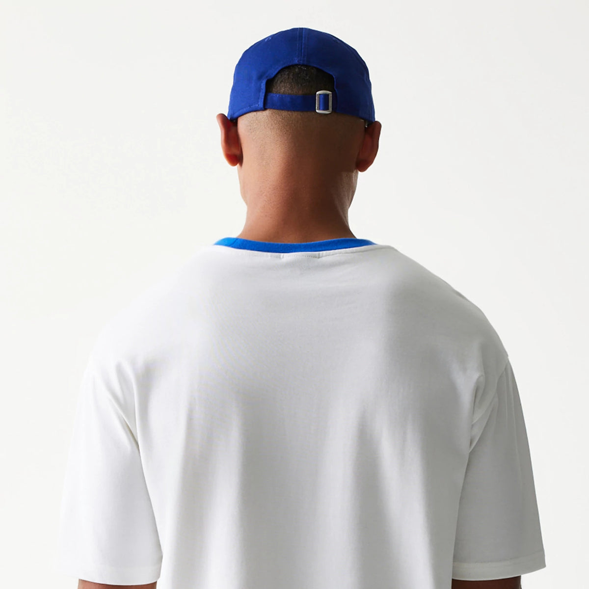 New Era Script White Oversized T-Shirt, back view on-model showing a plain white back with a contrasting blue collar, styled with a blue cap featuring an adjustable strap at the back.