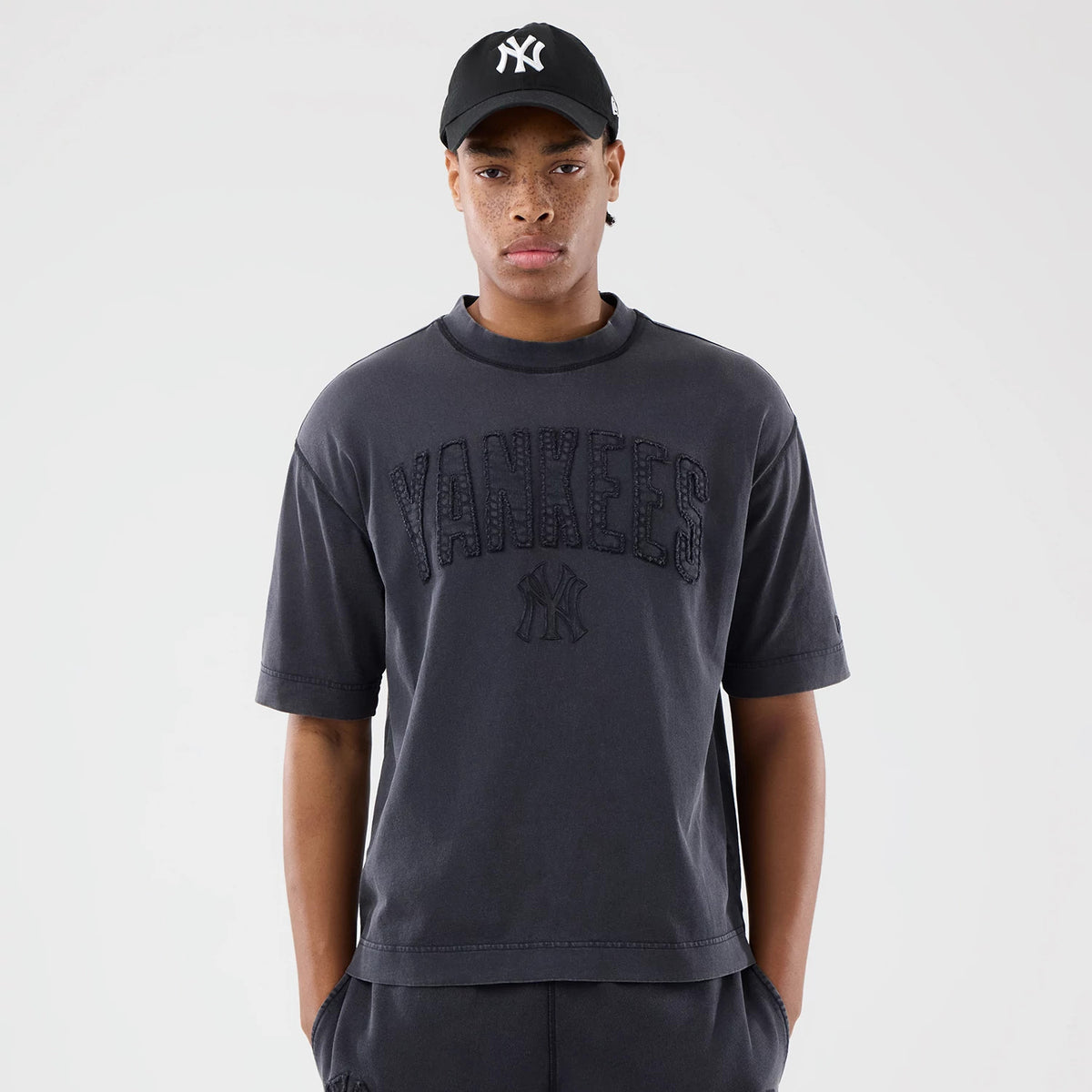 New Era New York Yankees MLB Washed Black T-Shirt, front view on-model featuring large embroidered Yankees lettering across the chest below a small Yankees logo, styled with a black cap with a raised white New Era logo.