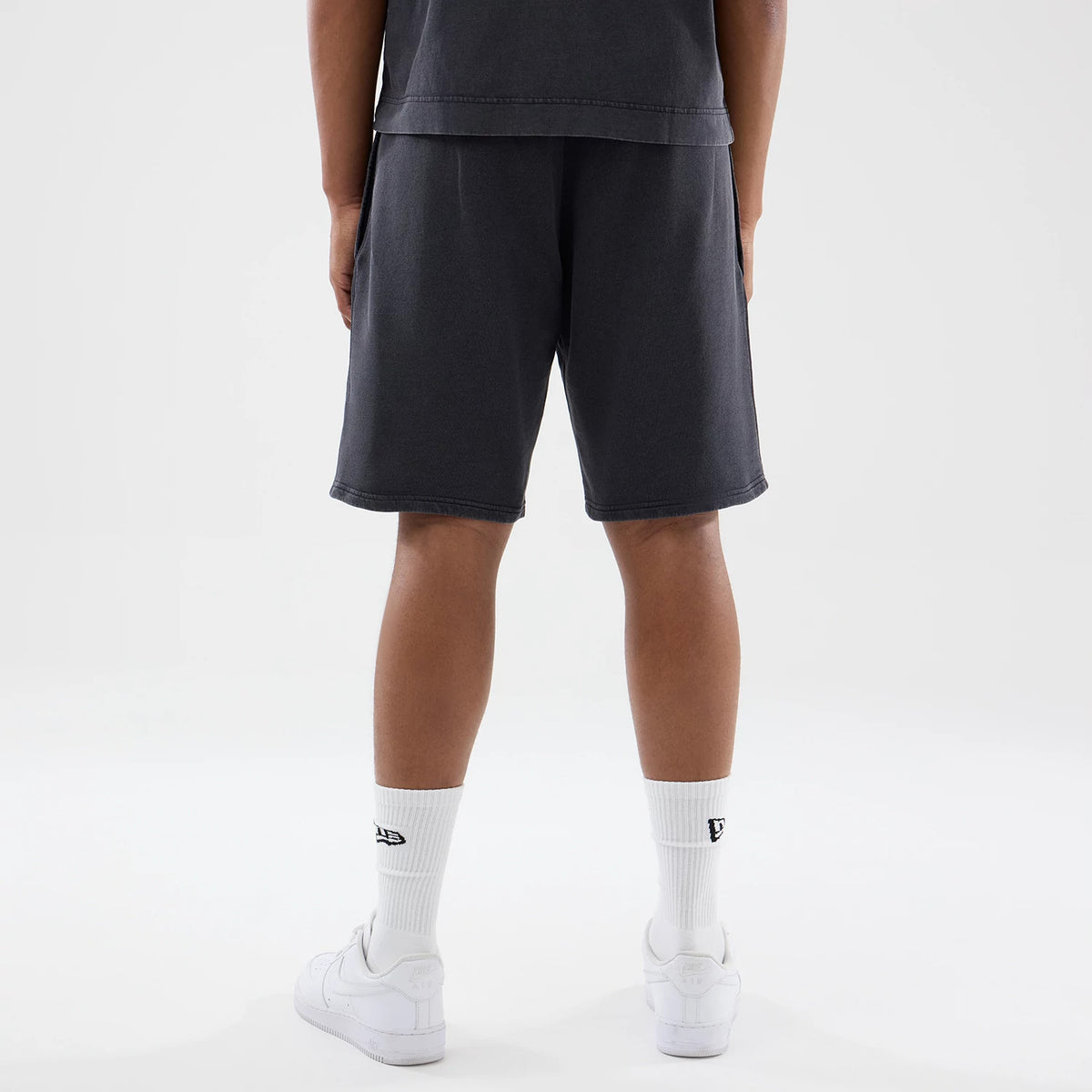 New Era New York Yankees MLB Washed Black Shorts, back view on a model showing the washed black fabric reaching above the knee, with plain hems and side pockets, styled with white socks and trainers.
