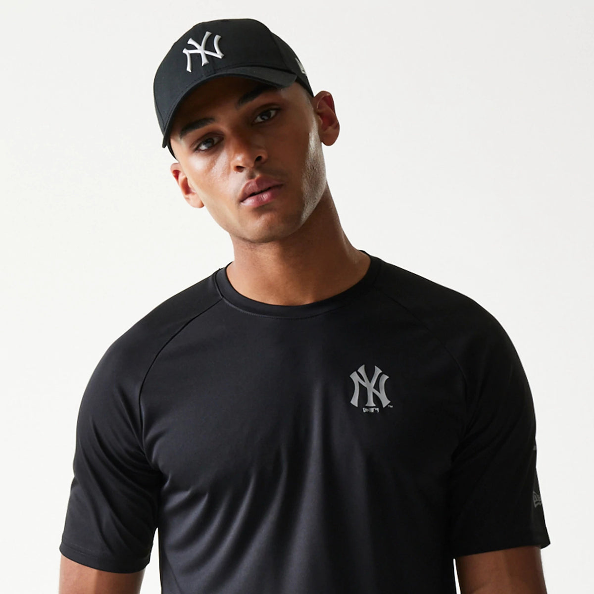 New Era New York Yankees MLB Reflective Black T-Shirt, front view on-model showing a reflective NY logo printed on the left chest, paired with a black cap featuring an embroidered white NY logo at the front.