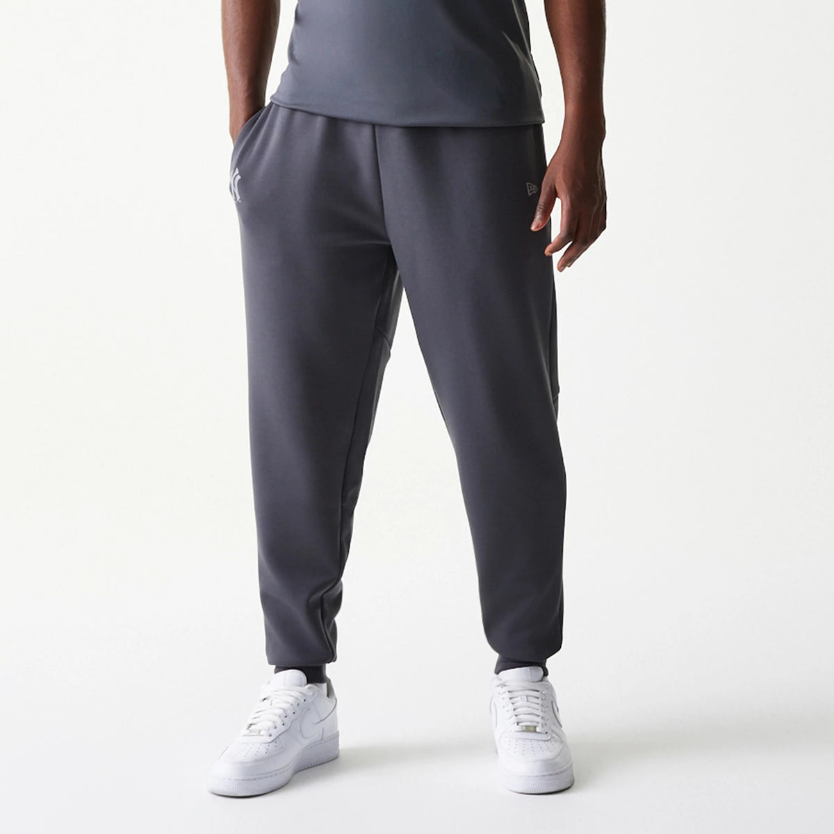 New Era New York Yankees MLB Dark Grey Joggers, front view on a model with hands in pockets, featuring the Yankees logo in white on the left thigh and the New Era flag logo on the right thigh above the cuffed ankles.